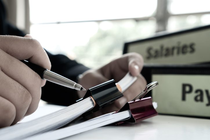 A person reviews documents with a pen in hand next to binders labeled Salaries and Payroll on a desk.