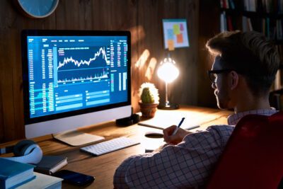 A person sits at a desk analyzing financial data and stock charts displayed on a large computer monitor in a dimly lit office.
