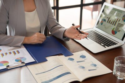 Person in business attire reviews charts and graphs on paper documents and a laptop at a desk, discussing financial data.