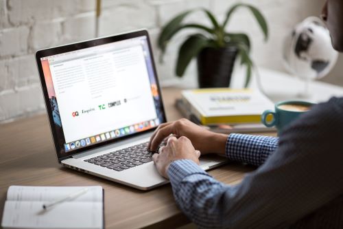 Person typing on a laptop at a wooden desk with an open notebook, books, a small globe, and a potted plant in the background.