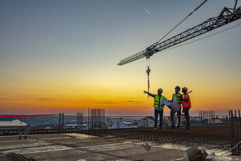 Three construction workers in safety vests and helmets stand on a building site at sunset, with a crane overhead, reviewing plans and pointing toward the horizon.