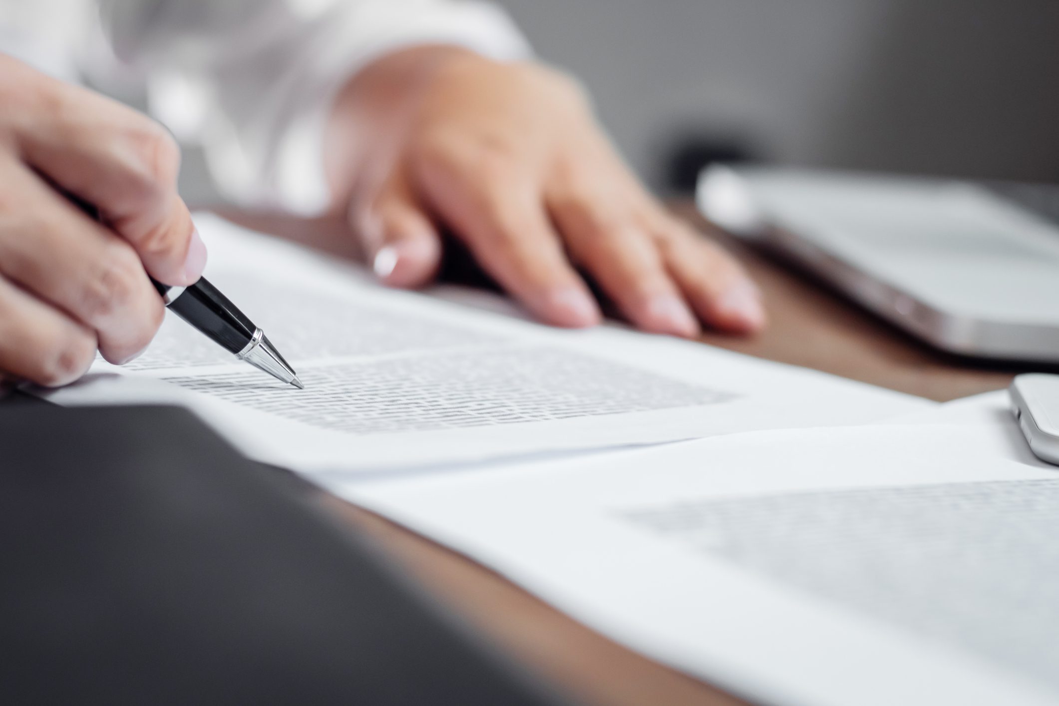 A person reviews and marks text on printed documents with a pen, with a laptop partially visible on the desk.