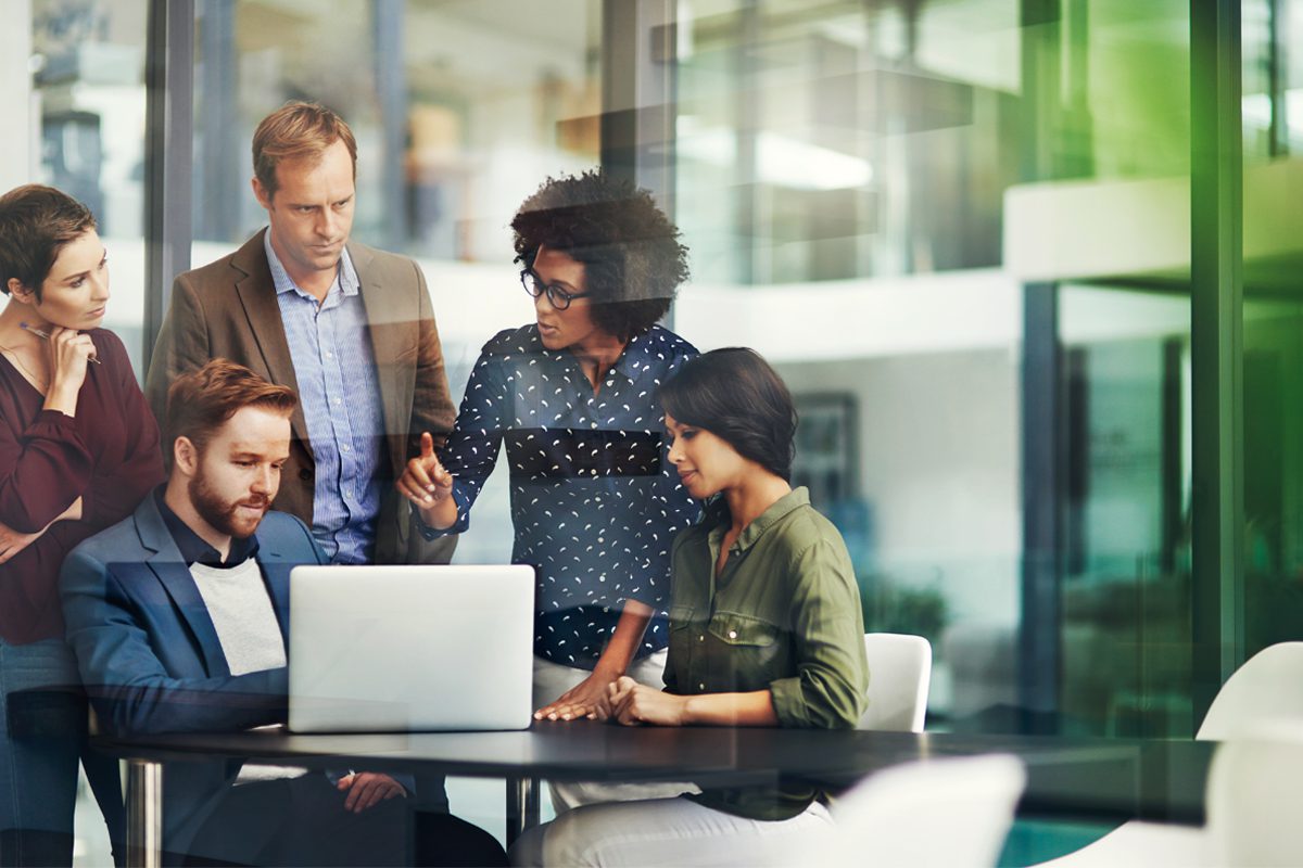 A group of five professionals gathers around a laptop in a modern office, discussing and collaborating on a project.