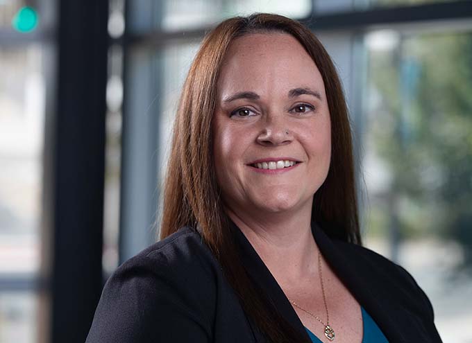 A woman with straight brown hair wearing a dark blazer and blue top smiles in front of a window with blurred background.