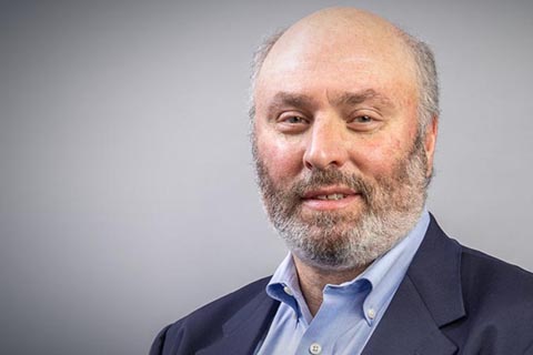 A middle-aged man with a beard and thinning hair wearing a blue blazer and light blue collared shirt, posing in front of a plain gray background.