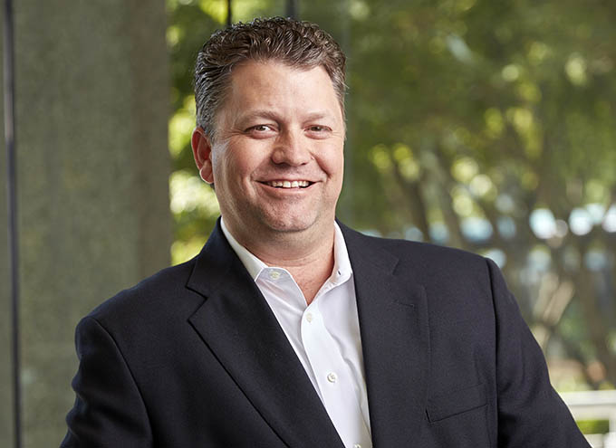 A man in a dark suit jacket and white shirt smiles while standing indoors with a blurred green outdoor background.