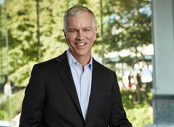 A man in a black suit and light blue shirt stands indoors near large windows, smiling, with trees and sunlight visible outside.