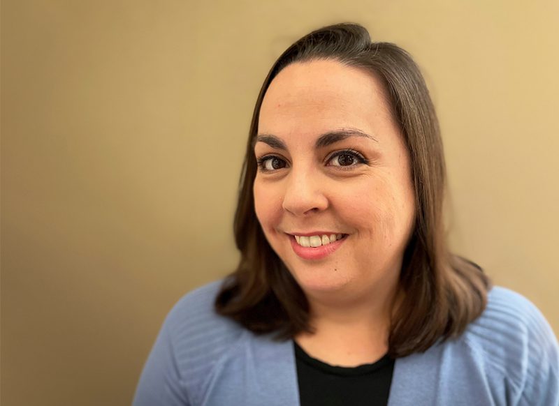 A woman with straight brown hair, wearing a light blue cardigan over a black top, smiles at the camera against a plain beige background.