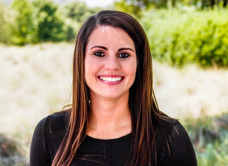 A woman with long brown hair wearing a black top is smiling outdoors with greenery and trees in the background.