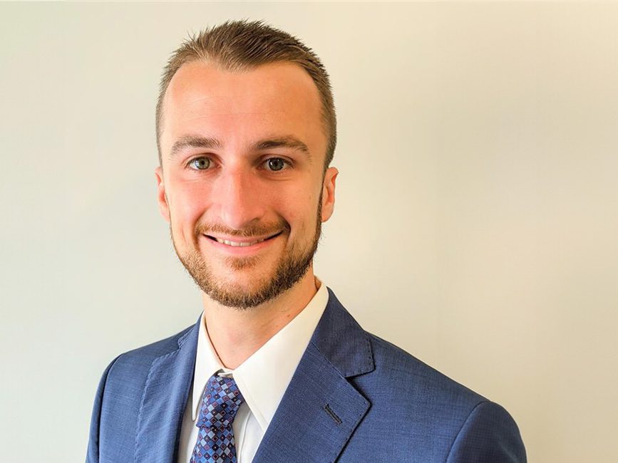 A man in a blue suit and patterned tie smiles at the camera against a plain light-colored background.