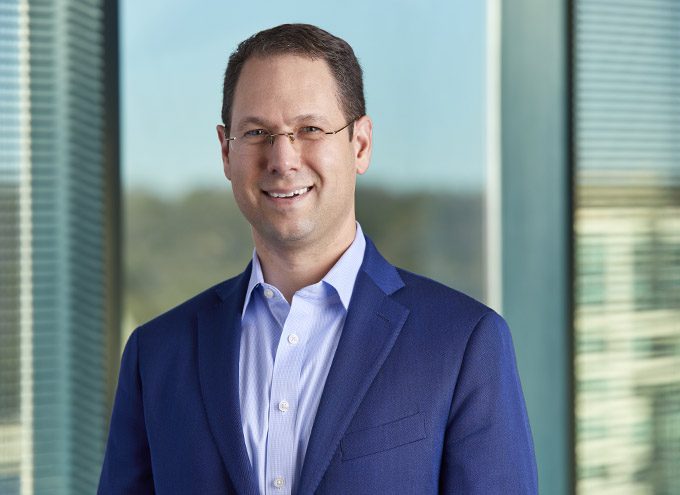 A man in a blue suit and glasses stands indoors in front of large windows, smiling at the camera.