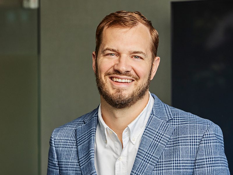 A man with short light brown hair and a beard, wearing a blue plaid blazer and white shirt, smiles while standing indoors.