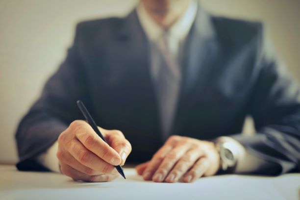 A person in a suit sits at a desk, writing on paper with a pen.