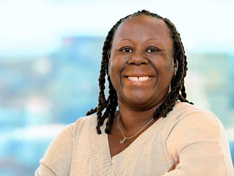 A woman with braided hair smiles, wearing a light sweater and a necklace, standing in front of a blurred background.