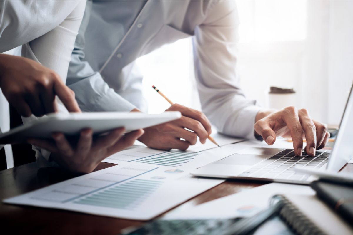 Two people review documents with charts, using a tablet and a laptop. One writes with a pencil, while the other points at the laptop screen.