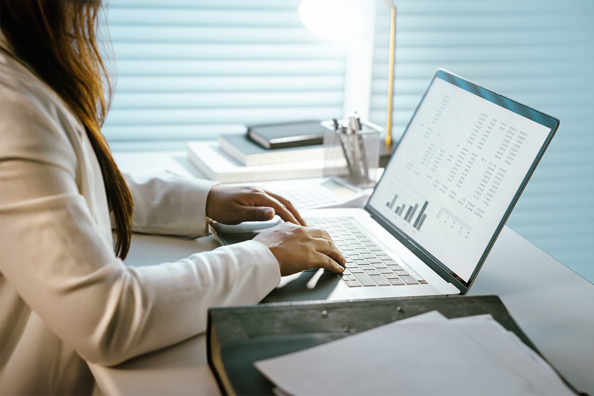 Person in a white blazer typing on a laptop displaying charts and data, sitting at a white desk with documents, a lamp, and office supplies.