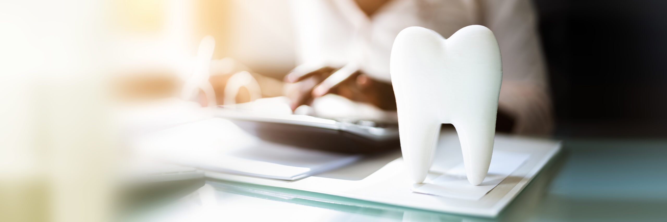 A large tooth model sits on a desk in the foreground, while a person works in the background with paperwork and a pen.