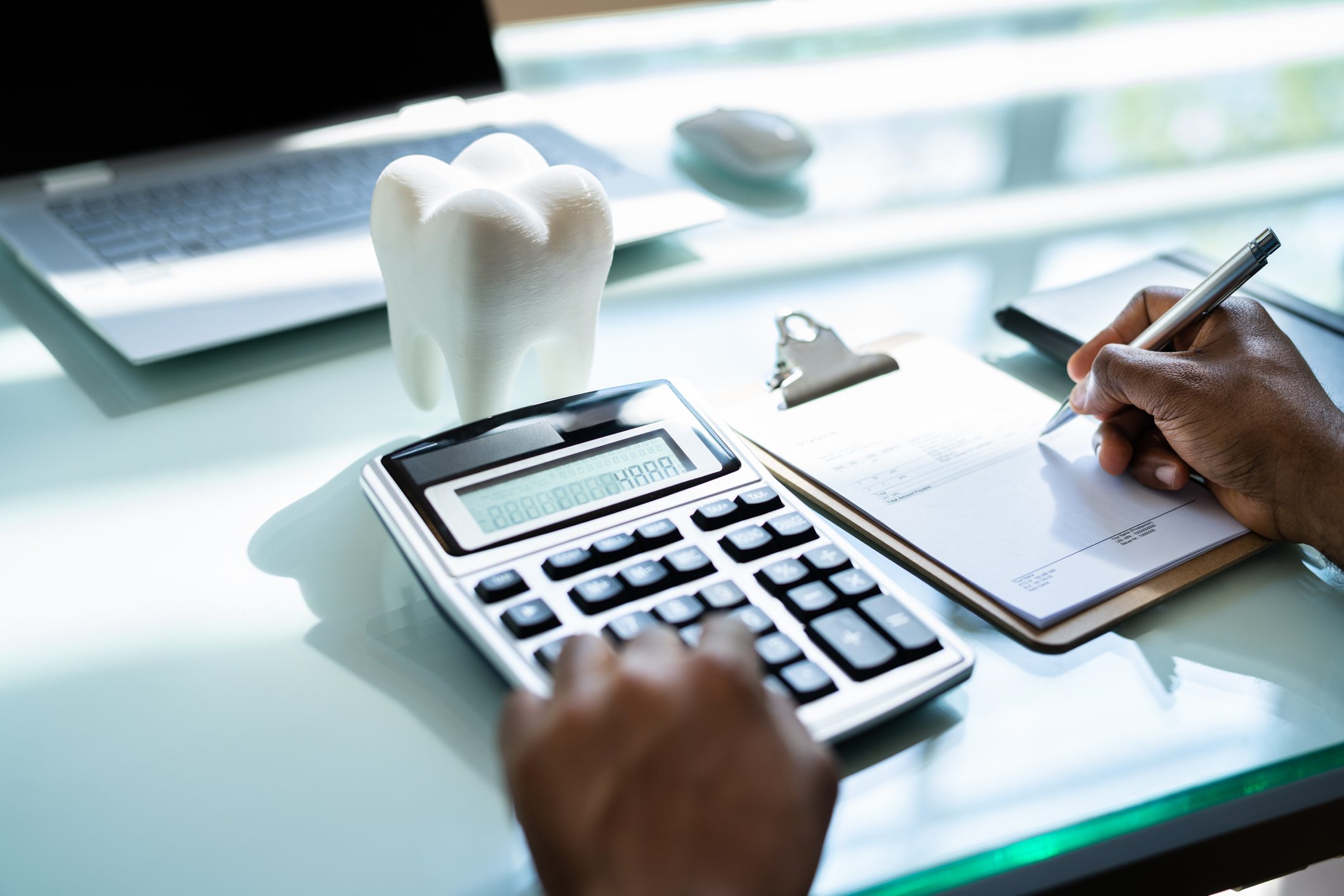 Person using a calculator and writing on a clipboard at a desk with a laptop and a model of a tooth.