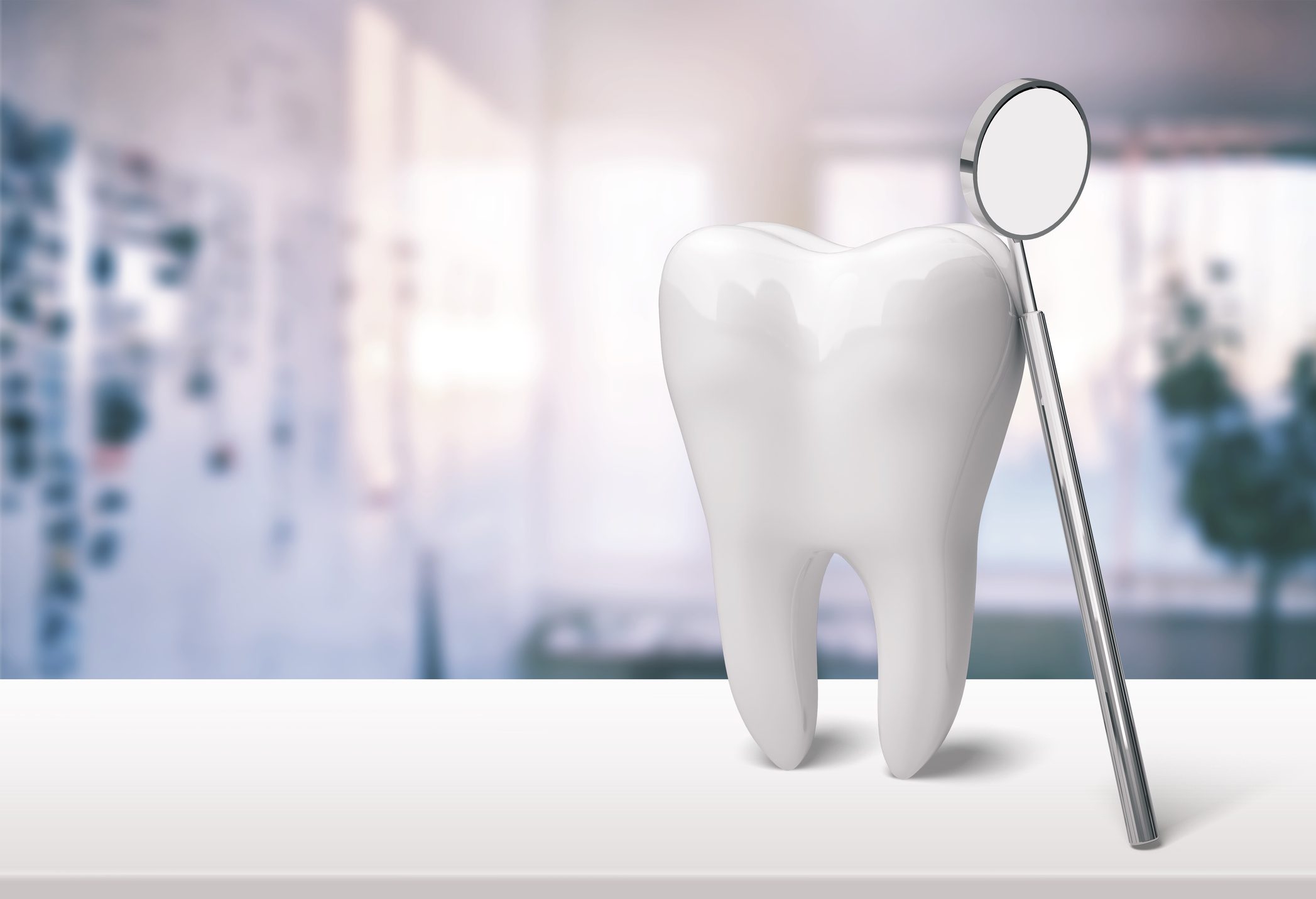 A model of a tooth stands upright next to a dental mirror on a white surface, with a blurred indoor background.
