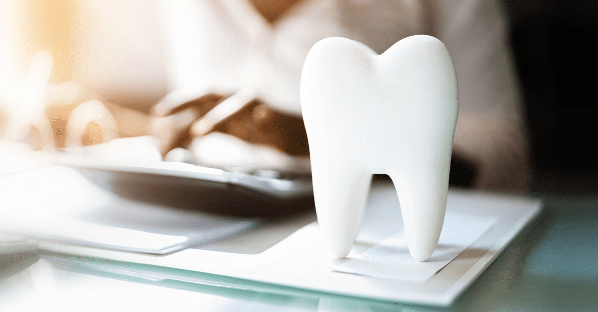 A large white tooth model sits on a desk with papers, while a person in the background works on a tablet.