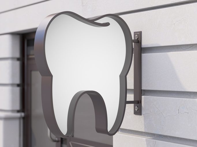 A metal sign in the shape of a tooth is mounted on the exterior wall of a building, indicating a dental clinic or dentist’s office.