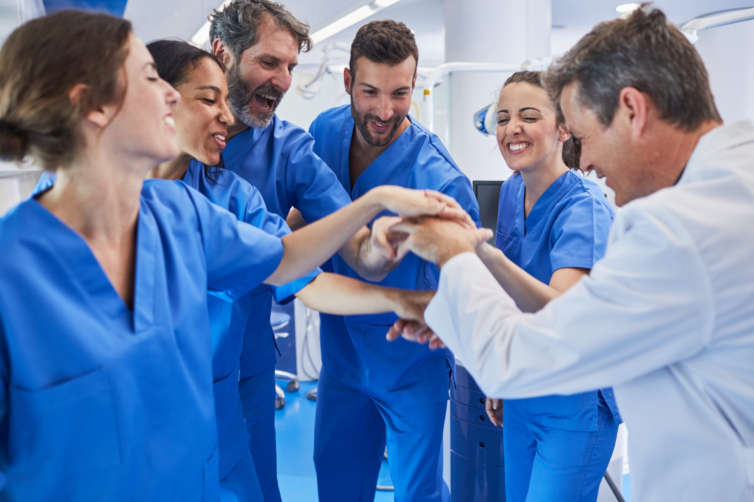 A group of healthcare professionals in blue scrubs stand together, smiling and placing their hands in the center as a gesture of teamwork.