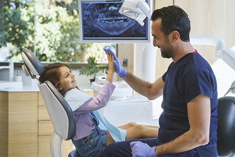 A young girl sitting in a dental chair gives a high-five to a dentist wearing gloves. Dental equipment and an x-ray image are visible in the background.