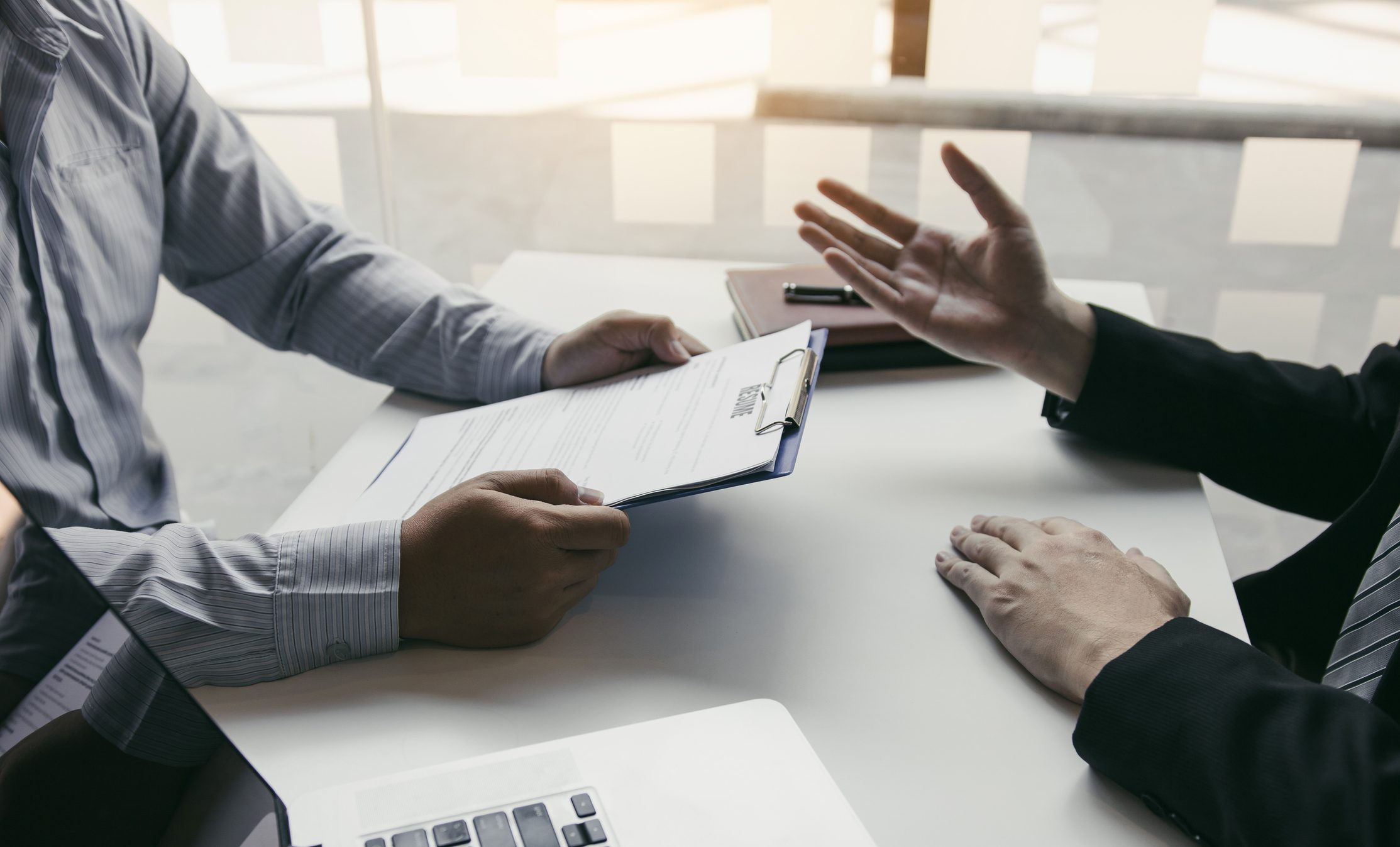 Two people sit at a desk discussing a document on a clipboard; one person is holding the clipboard while the other gestures with their hand.