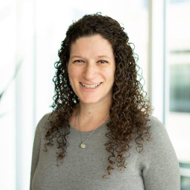 A woman with long curly hair, wearing a gray sweater and a necklace, stands indoors in front of large windows, smiling at the camera.