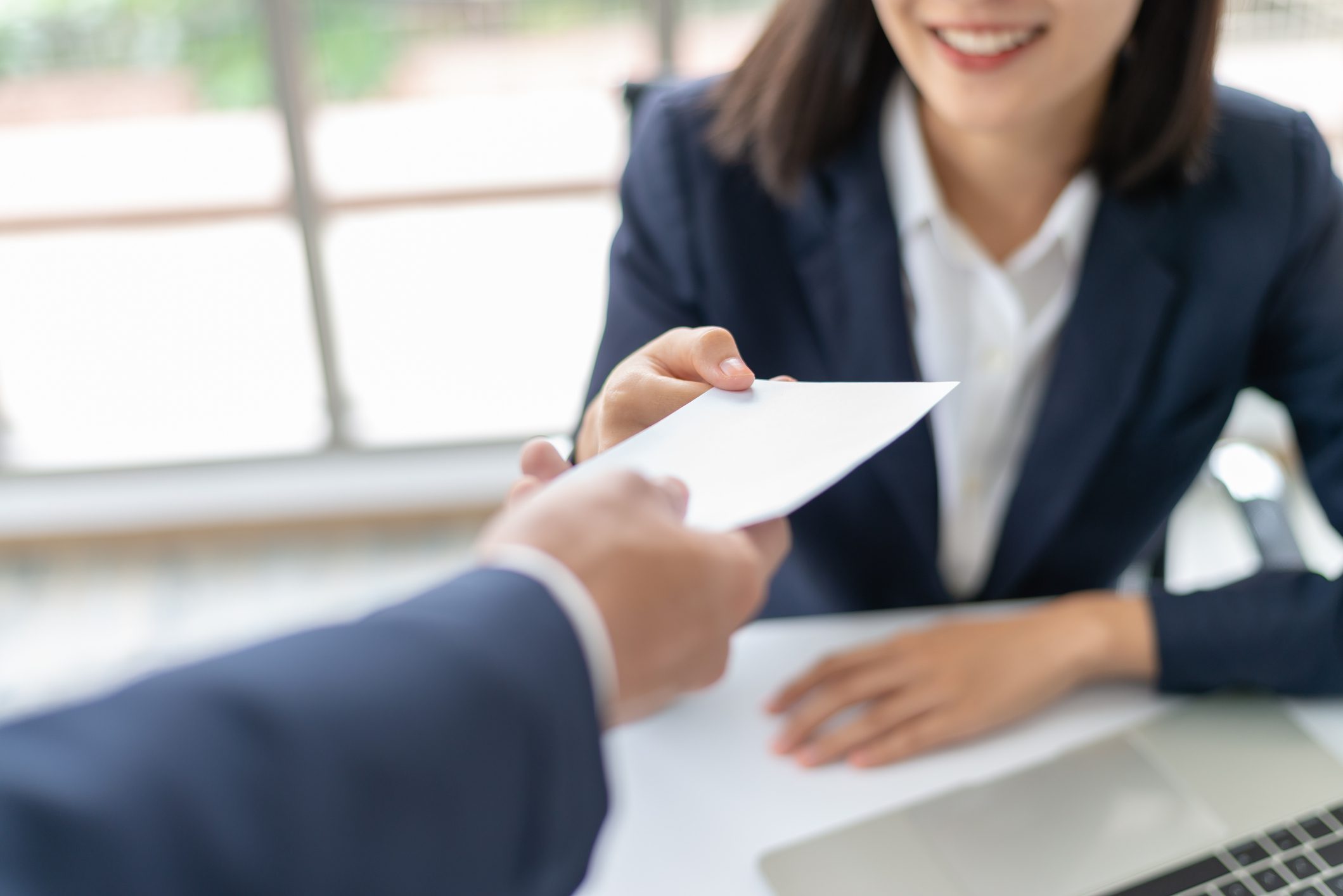 A person in a suit hands an envelope to a smiling woman in business attire at a desk, with a laptop nearby.