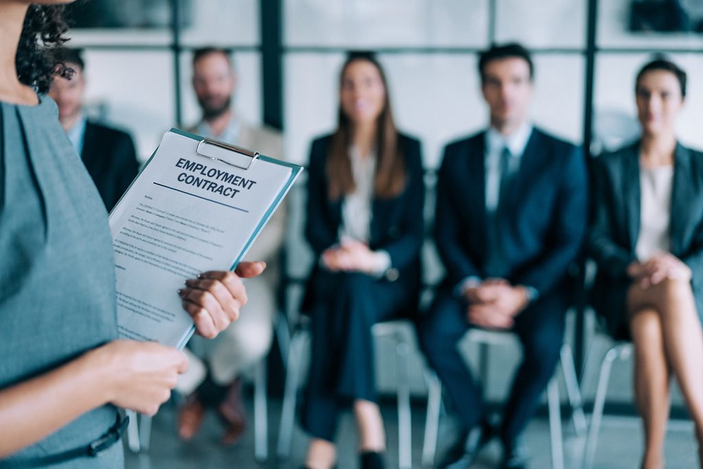 A person holding an employment contract on a clipboard stands in front of four seated individuals in formal business attire.