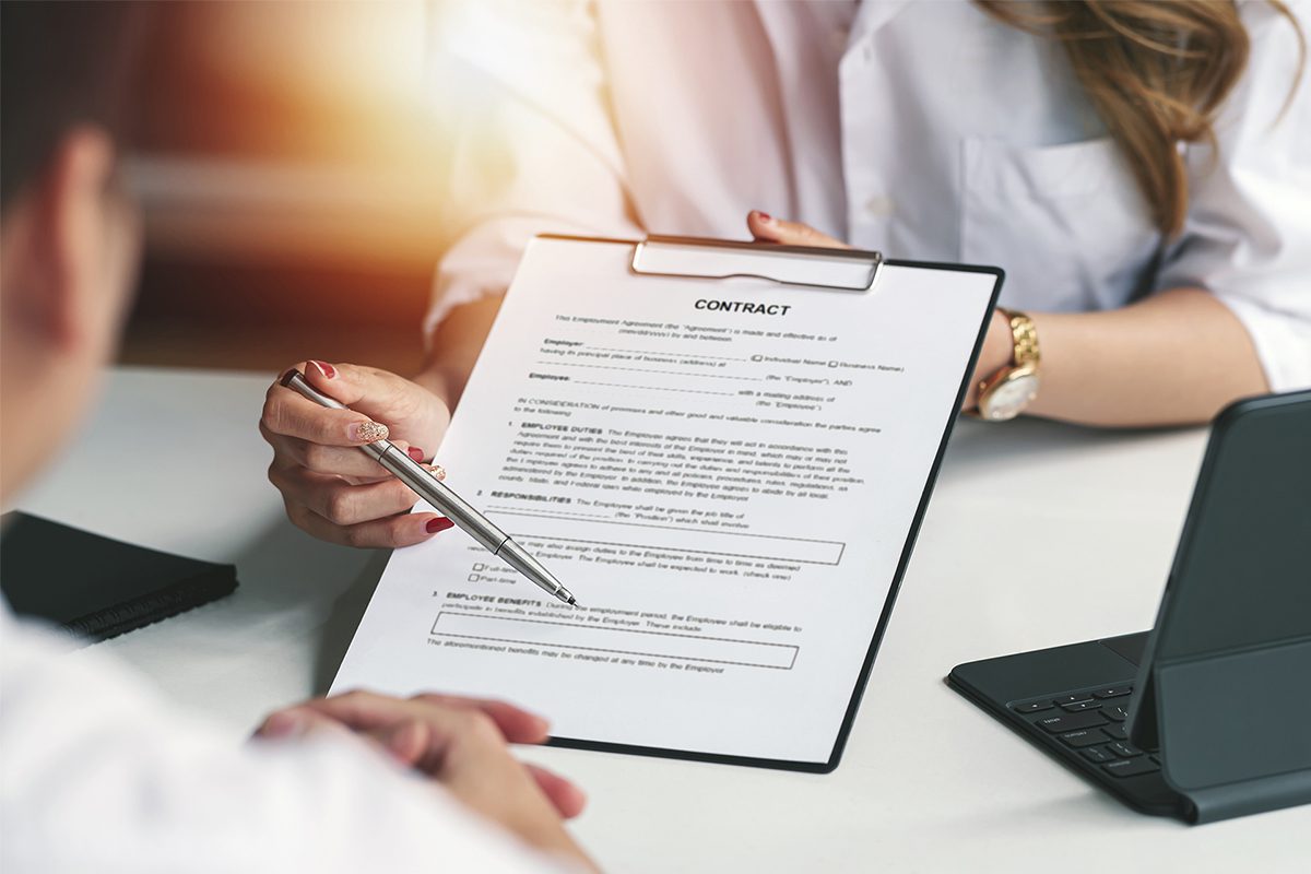 A person holding a clipboard with a contract, pointing at the document with a pen during a meeting with another individual at a desk.