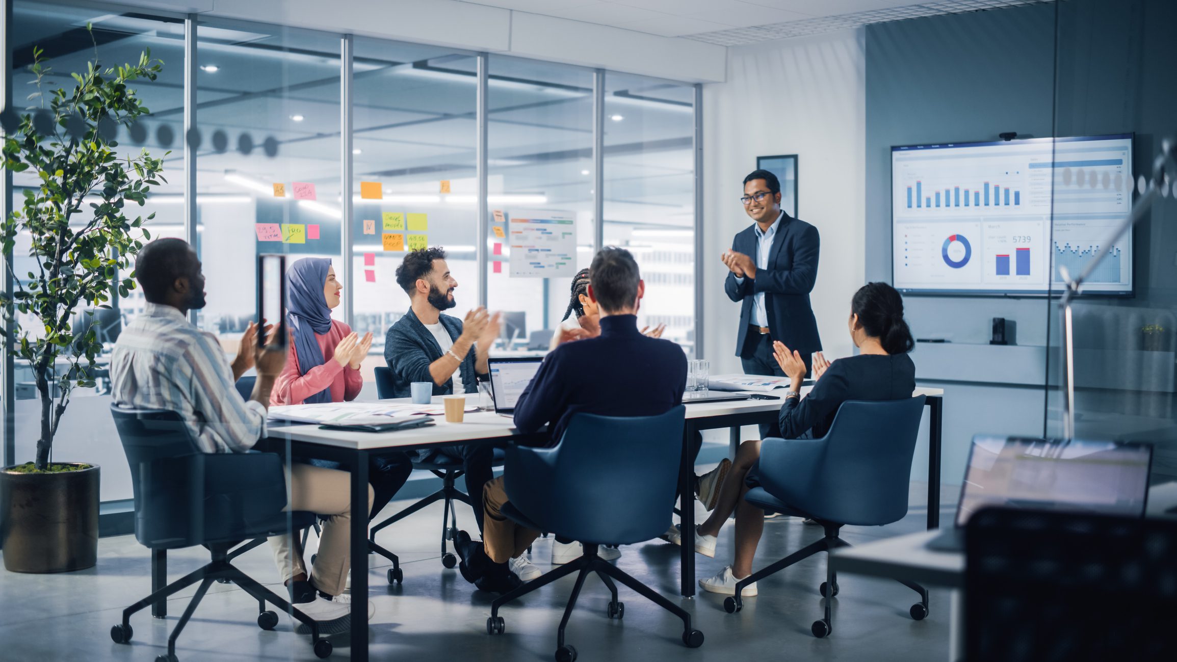 A group of professionals sit around a conference table, listening and applauding as a man in a suit presents data on a large screen in a modern office setting.