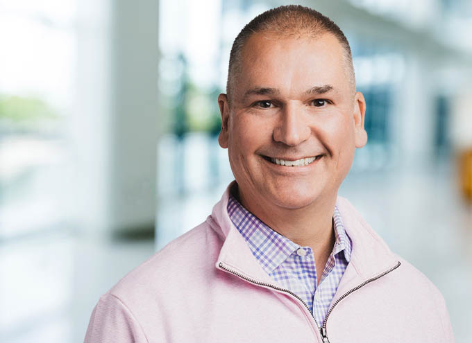 Smiling man in a light pink quarter-zip sweater and checkered shirt, standing in a bright, modern indoor setting.
