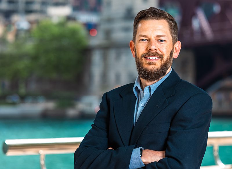 A man with a beard wearing a dark blazer and blue shirt stands outdoors with his arms crossed in front of a river and cityscape background.