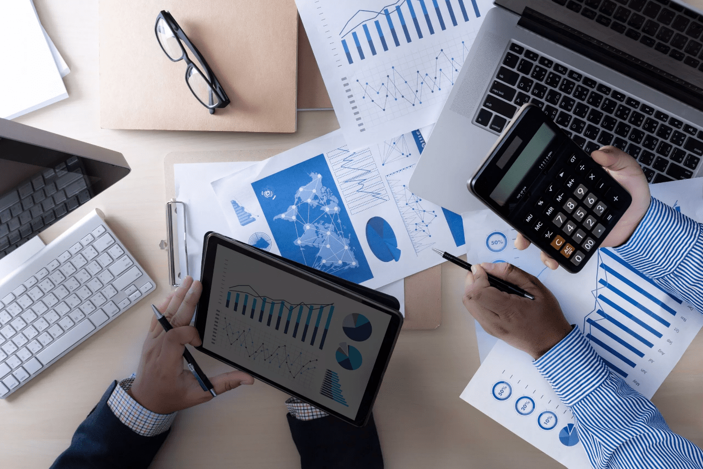 Two people analyze financial charts and graphs on paper, a tablet, and a laptop, with one person using a calculator at a cluttered office desk.