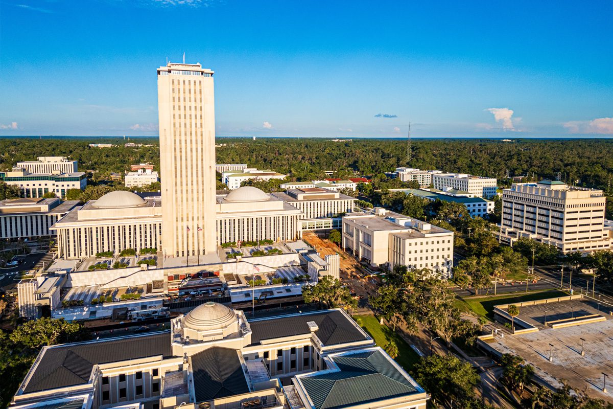 Aerial view of a large government complex with a tall central tower, domed buildings, and surrounding trees under a clear blue sky.