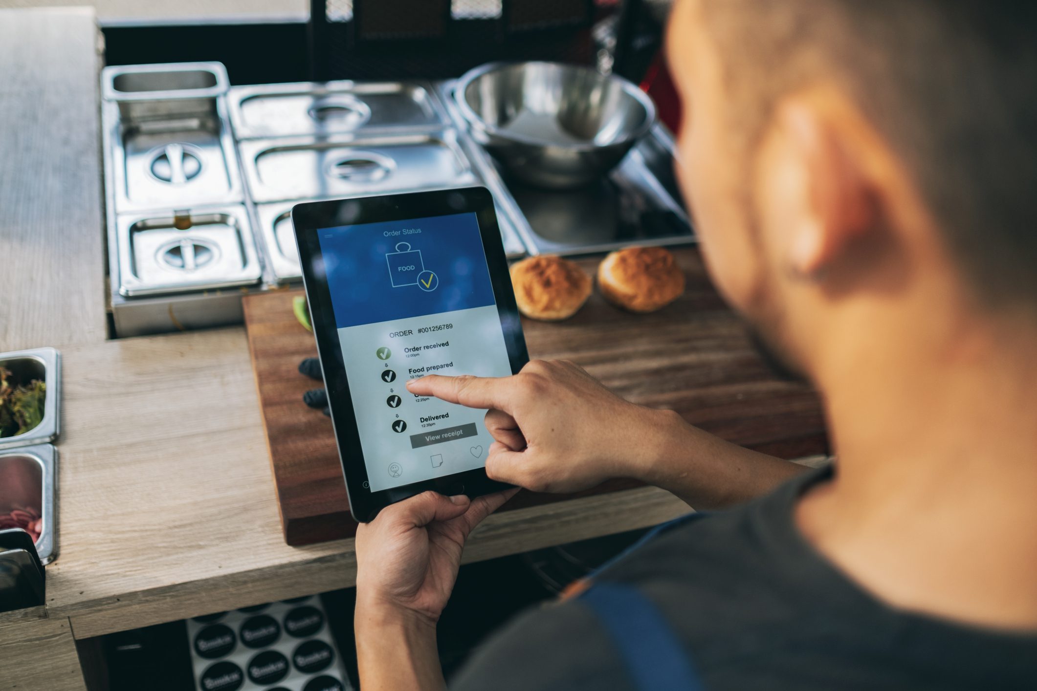 A person uses a tablet to check order status in a kitchen setting, with ingredients and prepared food visible on the counter.