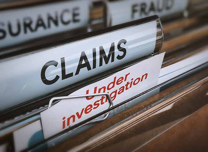 A close-up of file folders labeled CLAIMS, FRAUD, and INSURANCE, with a document titled Under investigation clipped inside the CLAIMS folder.