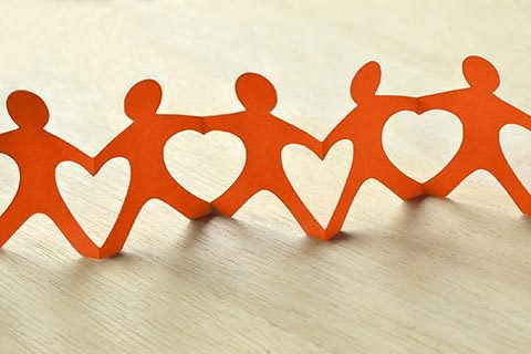 A row of orange paper cutout figures holding hands, each with a heart-shaped cutout in the center, placed on a light wooden surface.