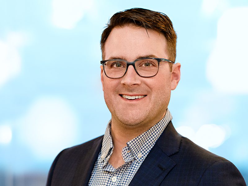 A man wearing glasses, a dark blazer, and a checked shirt smiles at the camera against a blurred blue background.