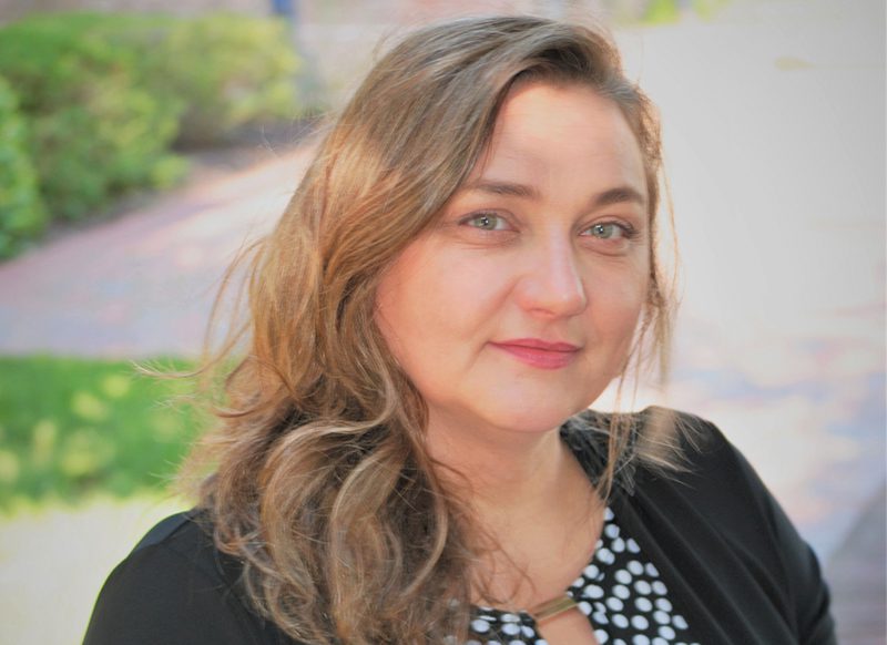 A woman with wavy light brown hair and blue eyes is wearing a black jacket over a black and white polka dot top, sitting outdoors with greenery in the background.