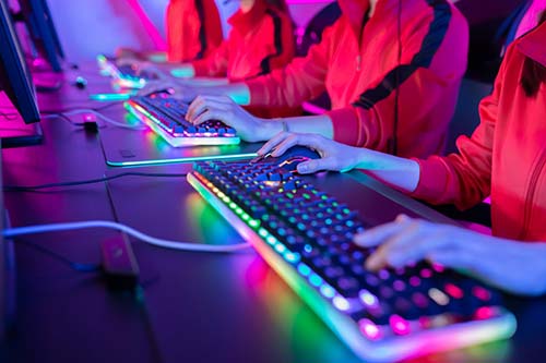 Three people in red jackets use illuminated RGB keyboards and monitors in a dark room, likely participating in a group gaming or esports session.