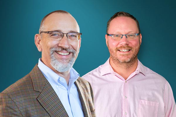 Two men smiling and facing the camera; one wears a checked blazer and glasses, the other wears a light pink shirt and glasses, against a teal background.