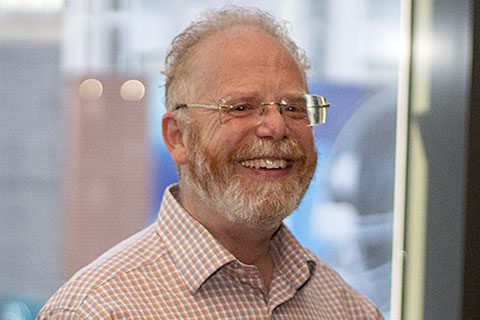A middle-aged man with glasses and a beard, wearing a plaid shirt, smiling indoors near a window.