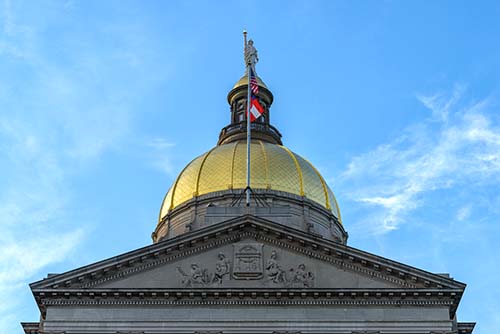 A gold-domed government building with an American flag and another flag on top, set against a blue sky with light clouds.