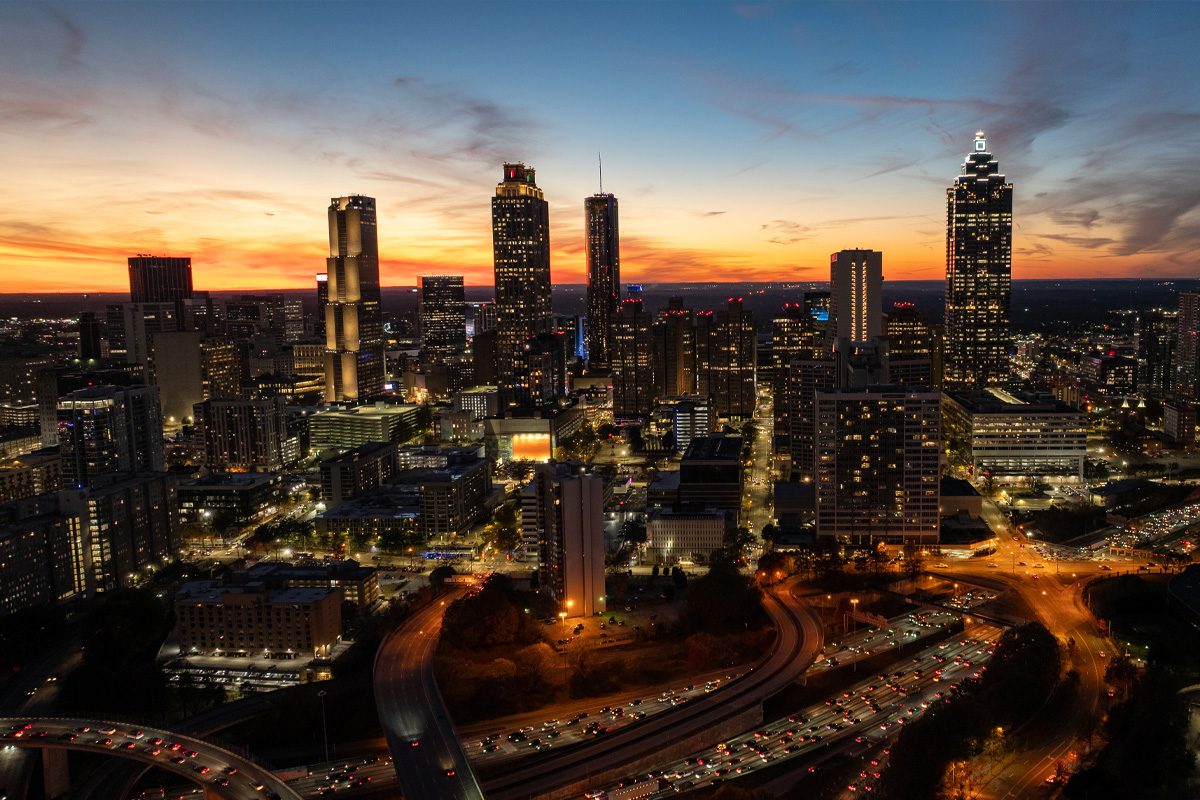 Aerial view of a city skyline at dusk with illuminated buildings, busy highways, and a colorful sunset in the background.