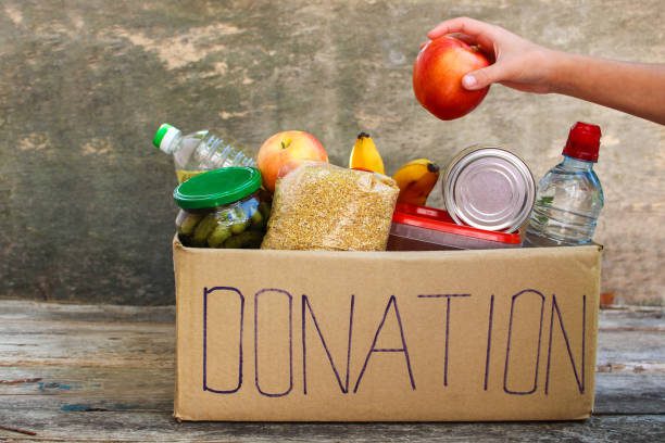 A cardboard box labeled DONATION filled with food items such as canned goods, fruit, grains, and bottled water; a hand places an apple inside.