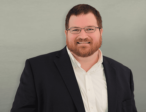 Man with short brown hair, beard, and glasses wearing a dark suit jacket and white shirt, standing and smiling in front of a plain light background.