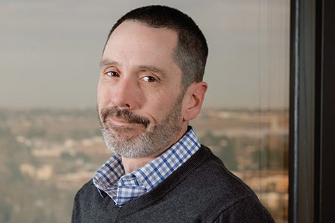 A man with short dark hair and a trimmed beard wearing a blue checkered shirt and dark sweater stands indoors in front of a window with an outdoor view.